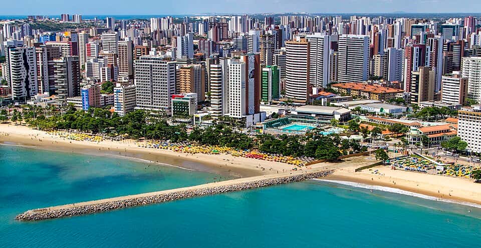 Orla de Fortaleza com praia de areia dourada, mar azul, coqueiros e skyline da cidade ao fundo.