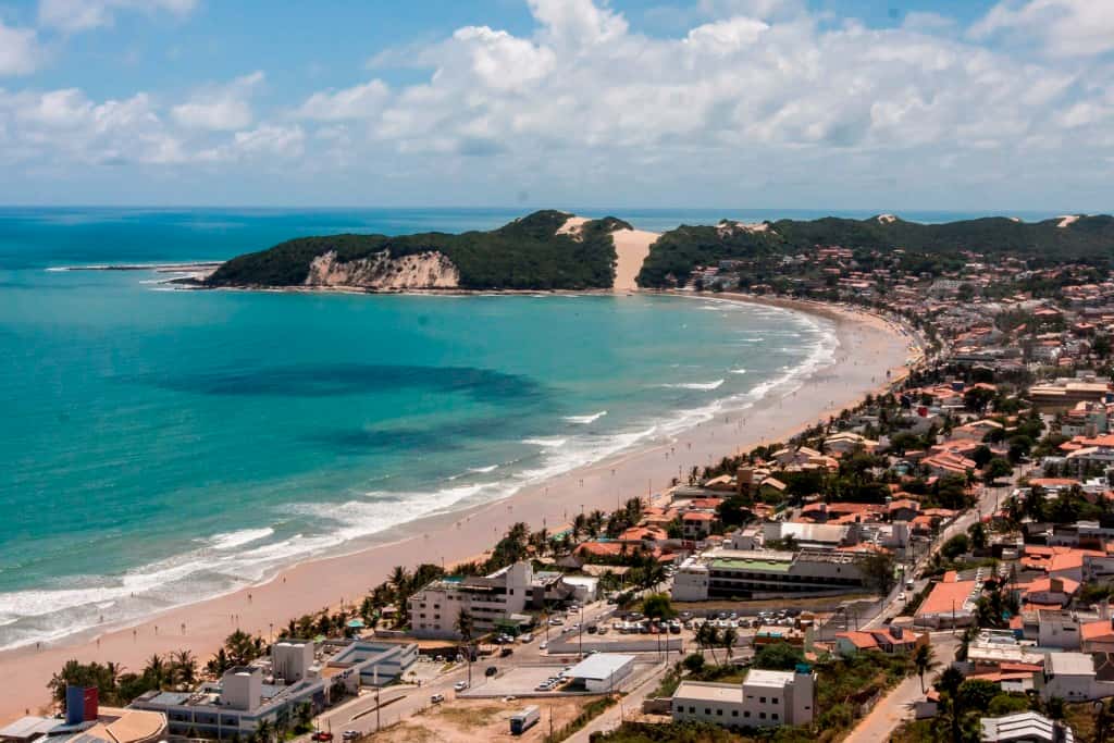 Praia de Ponta Negra em Natal com areia dourada, mar azul, coqueiros e o Morro do Careca ao fundo.
